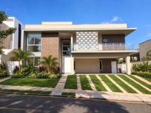 Fachada de residência contemporânea com cobogó branco, revestimento em tom terroso, grandes aberturas de vidro, varanda e paisagismo frontal.