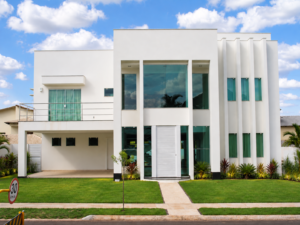 Fachada de residência contemporânea com volumes brancos, grandes aberturas de vidro, varanda lateral, elementos verticais e paisagismo frontal.