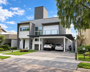 Fachada de residência contemporânea com volumes em tons de cinza, varanda com guarda-corpo de vidro, garagem coberta e jardim frontal.